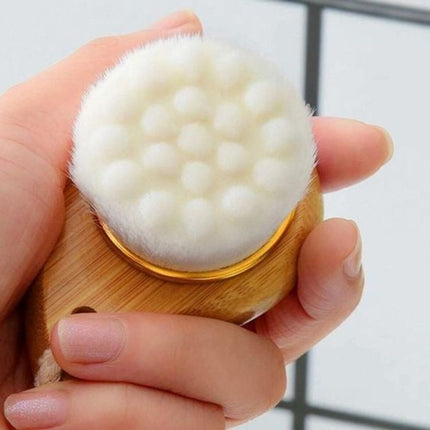 Hand holding a wooden facial cleansing brush with soft white bristle head against a neutral background